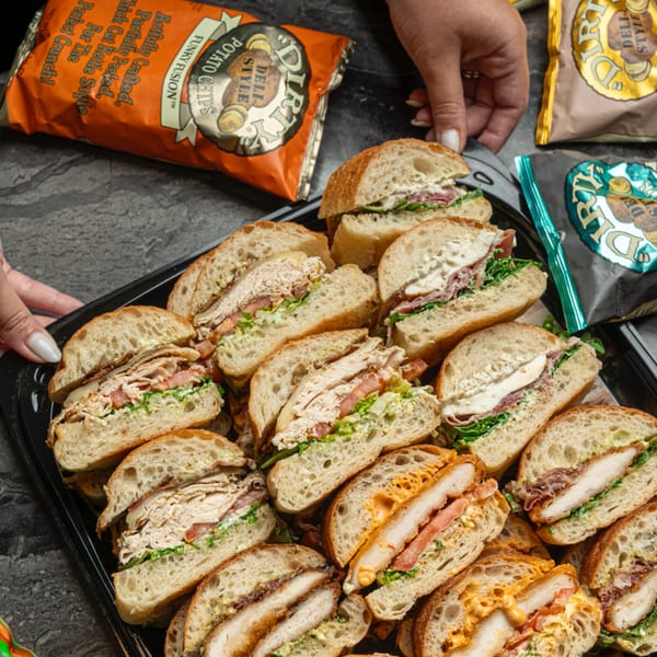 Hands serving a catering tray of various sandwiches with bags of Dirty Potato Chips.