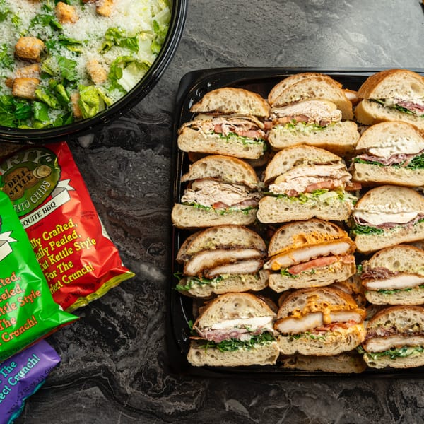 A catering tray of SNDWCH sandwiches, a Caesar salad, and bags of potato chips.