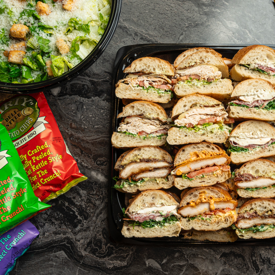 A catering tray of SNDWCH sandwiches, a Caesar salad, and bags of potato chips.