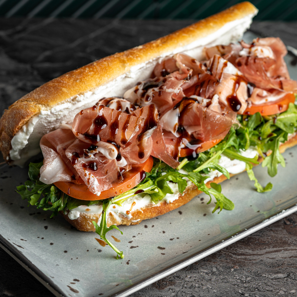 SNDWCH prosciutto sandwich with arugula, tomatoes, and balsamic glaze on a baguette.