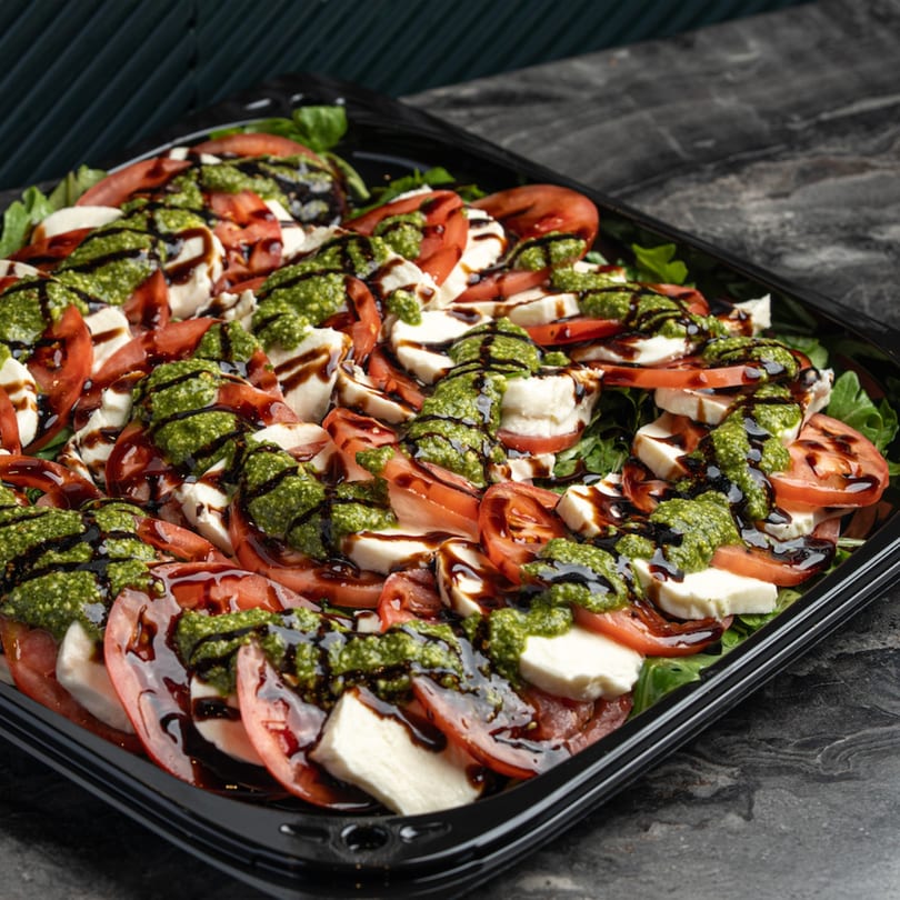 Caprese salad with sliced tomatoes, mozzarella, pesto, and balsamic glaze in a black tray.